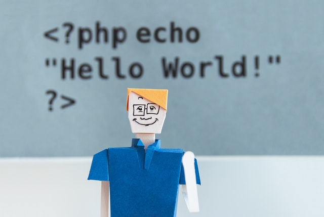 Cardboard man with glasses in front of some code, which says Hello World